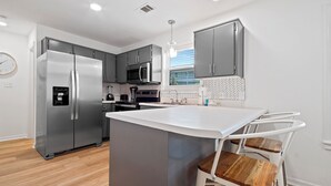 A modern kitchen with grey cabinets, a stainless steel refrigerator, a microwave, and a built-in oven. There is a white island with a sink and a chair with a wooden seat. - NEW LISTING The Proper Pelican 30A: STEPS to the BEACH! (Santa Rosa Beach)