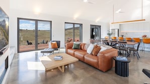 A modern living room with a leather sofa, a wooden coffee table, and a dining area with orange chairs. - Camber Farmhouse Jindabyne Countryside (Jindabyne)