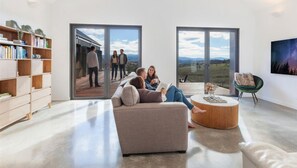 A modern living room with a sofa, a round coffee table, a bookshelf, and large sliding glass doors offering a view of a deck and mountains.