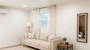A neutral-toned living room with a beige sofa, a small wooden side table, a lamp, and a mirror on the wall. - Private 1BR GuestHouse Near Charlotte Pet Friendly (Indian Trail)