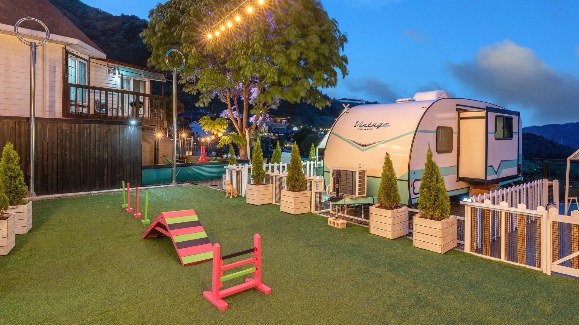 A backyard with artificial grass, a swing set, a playhouse, and a camper trailer.