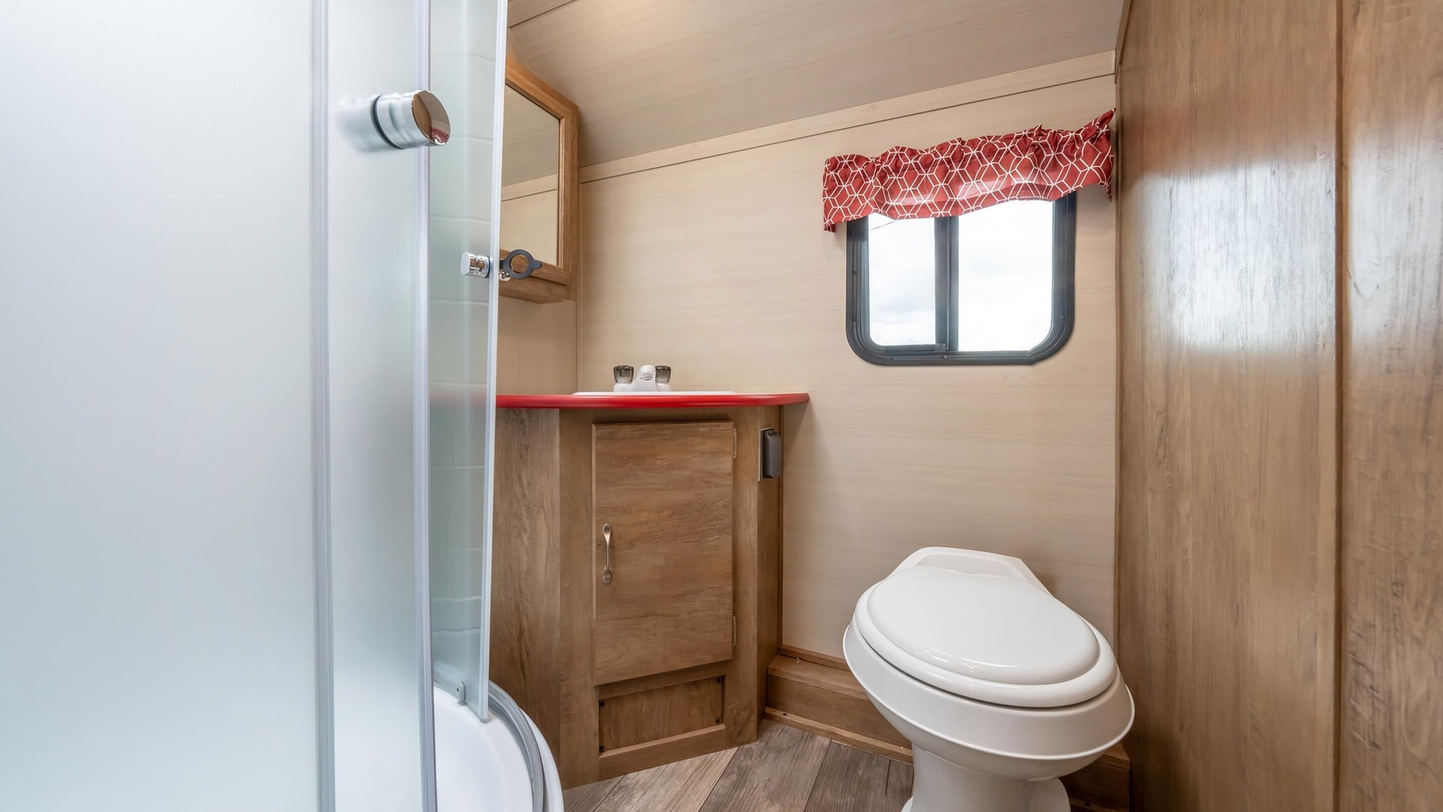 Ein kleines Badezimmer mit einem Holzkosmetikschrank, einer Toilette und einem kleinen Fenster mit einem roten, gemusterten Vorhang.