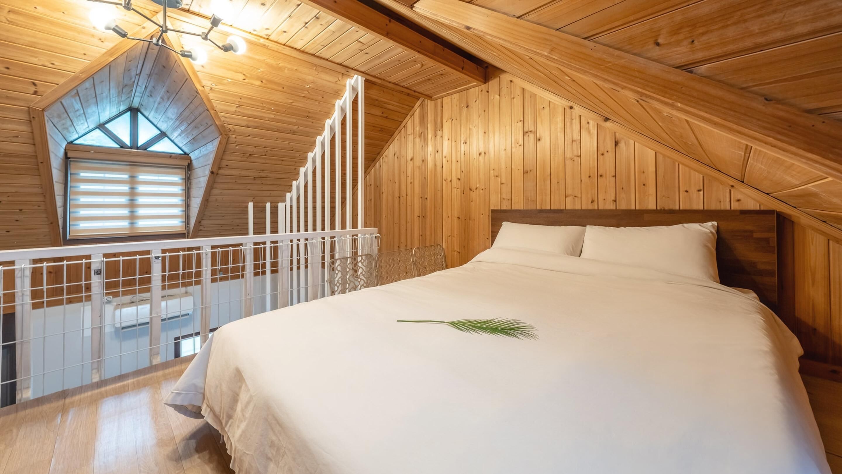 A bedroom with a wooden sloped ceiling, a large bed, and a window with blinds.