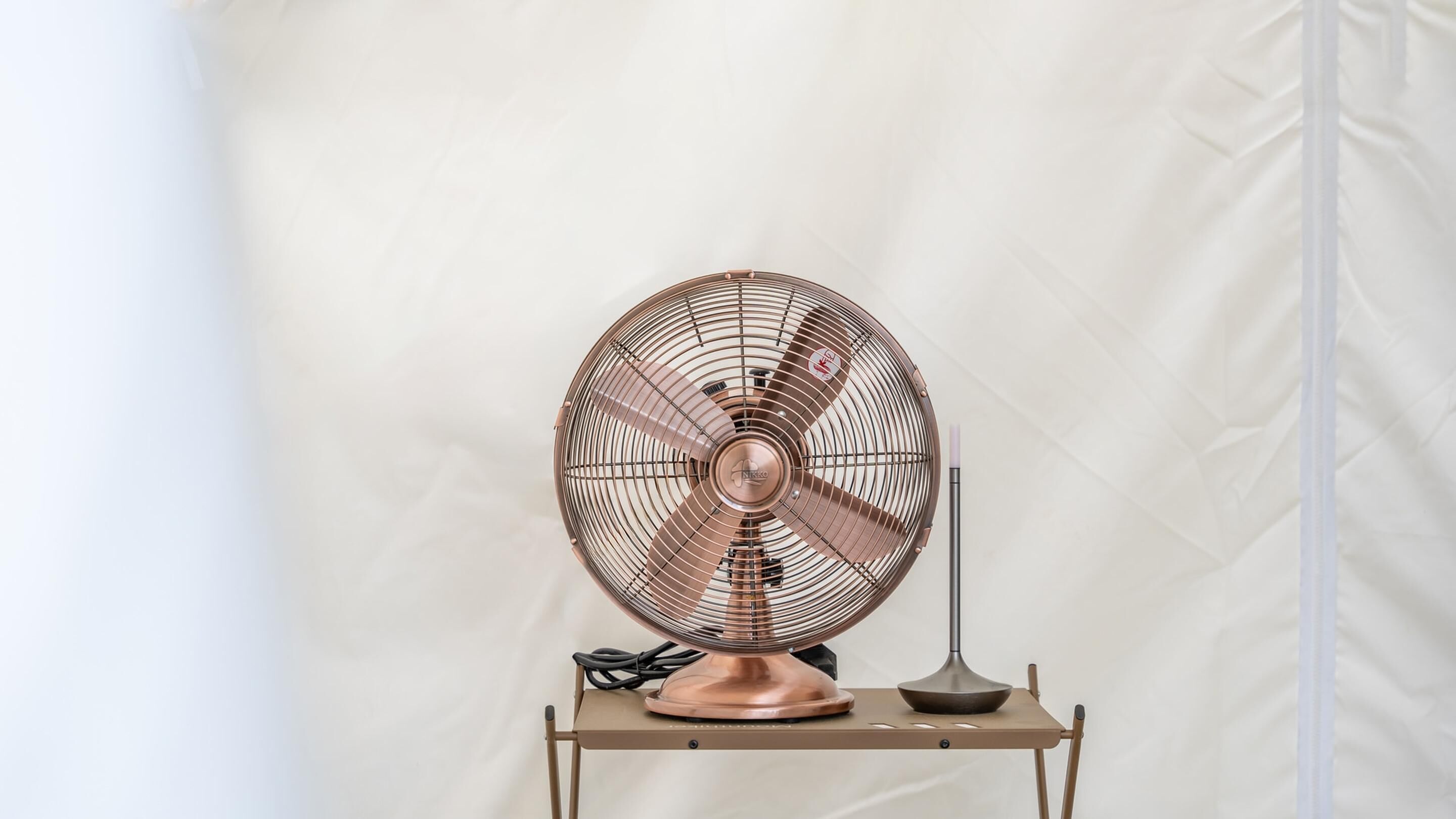 Un ventilador de pie con base de cobre y rejilla metálica, sobre una mesita con un control remoto a su lado.
