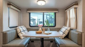 A compact RV interior with a dining table, cushioned seating, a window with a view, and a table lamp. - Gapyeong Dongban84 Glamping&pool Villa (Gapyeong)