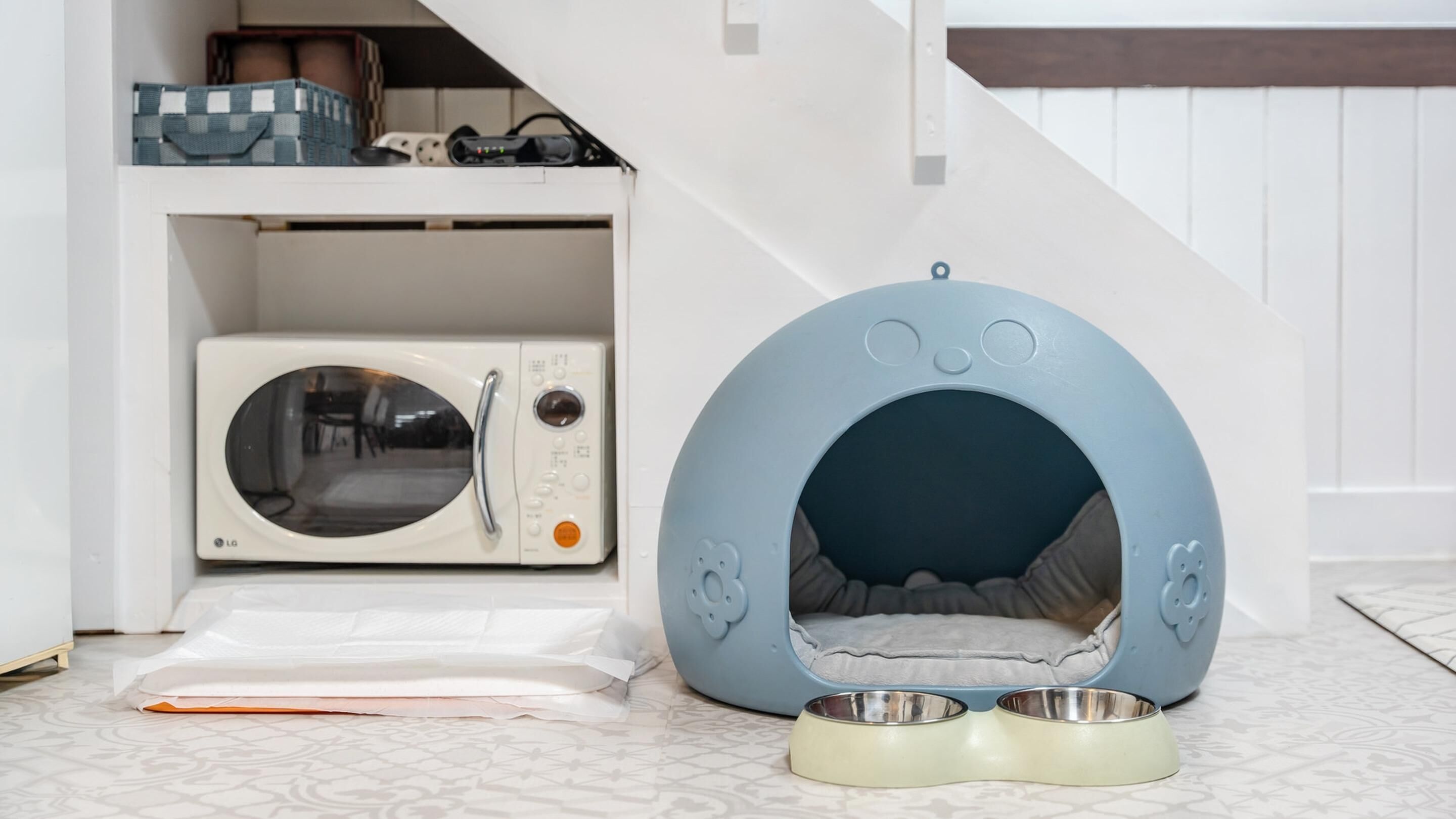 A pet bed with a blue exterior and a white interior, placed next to a microwave and a pet food bowl on a patterned floor.
