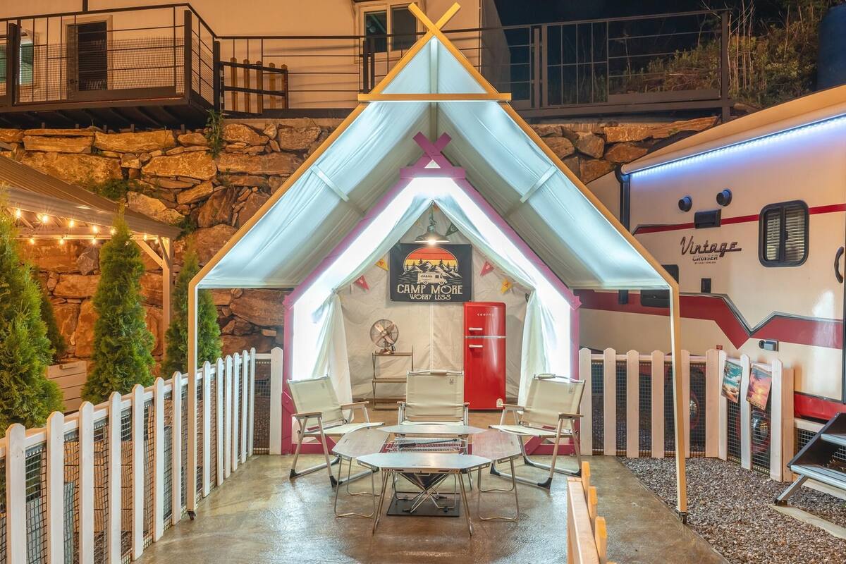 Un área de estar al aire libre acogedora, con una mesa y sillas, un refrigerador rojo y una caravana estacionada cerca.