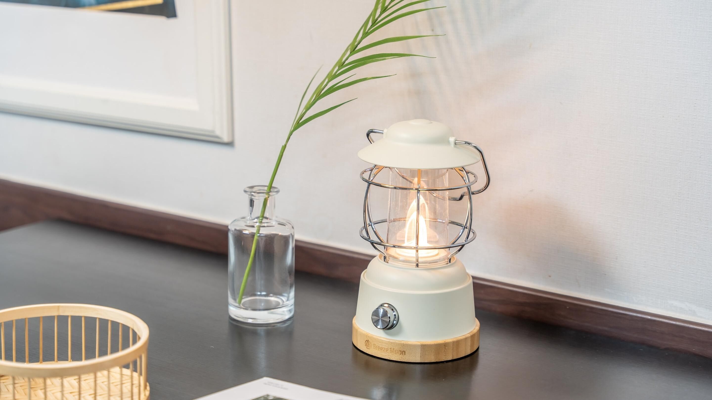 Una lámpara de mesa blanca con base de madera y pantalla de vidrio transparente, sobre una mesa de madera junto a un florero de vidrio con una planta verde.