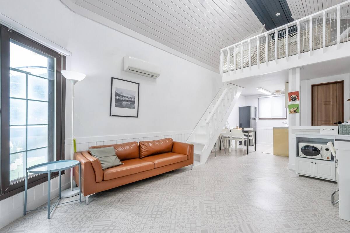 Una sala de estar moderna con un sofá de cuero marrón, un comedor, una cocina con microondas y un balcón con barandilla.