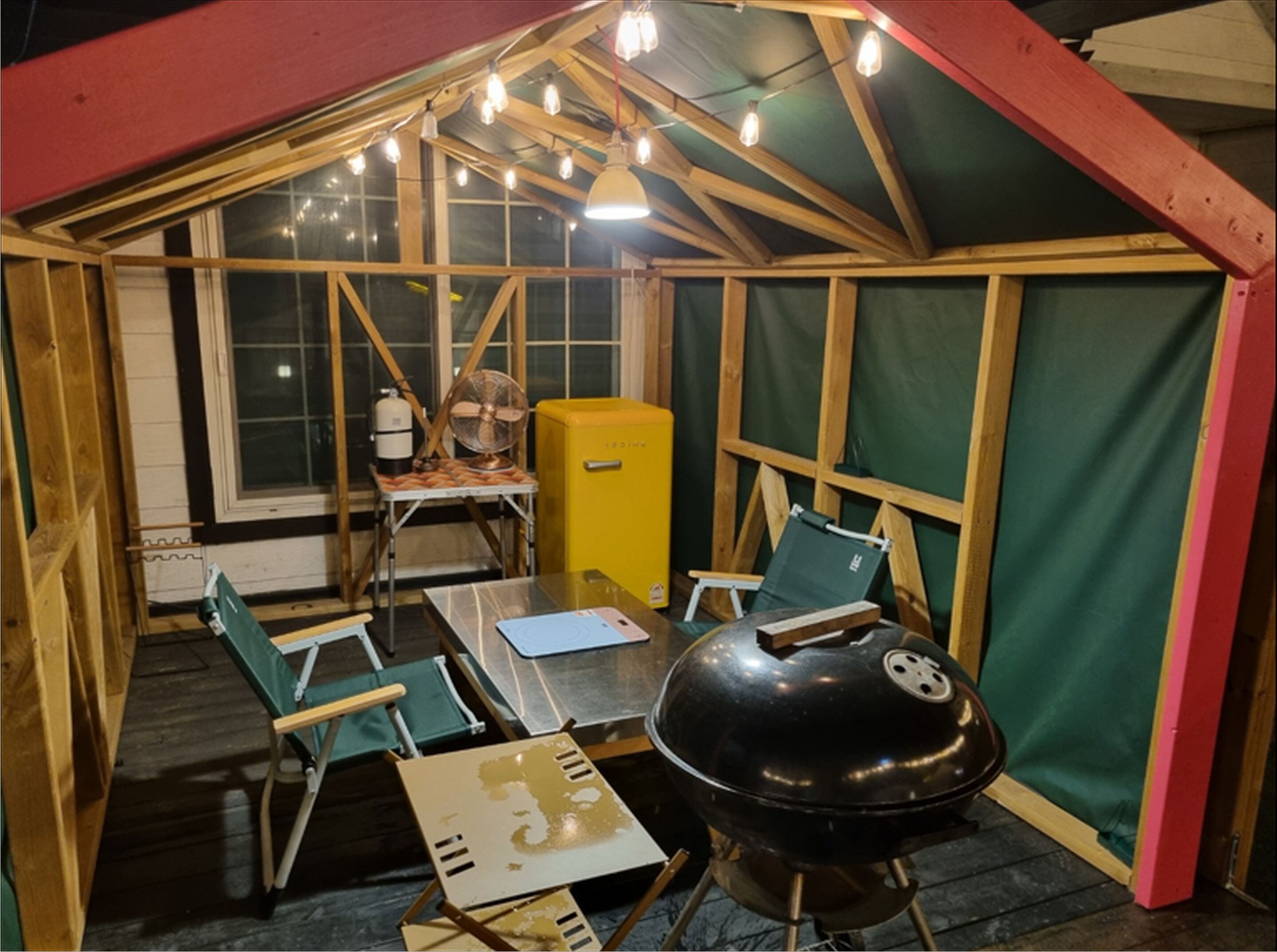 Un espacio al aire libre techado con una mesa, sillas, una parrilla, un ventilador y un electrodoméstico amarillo.