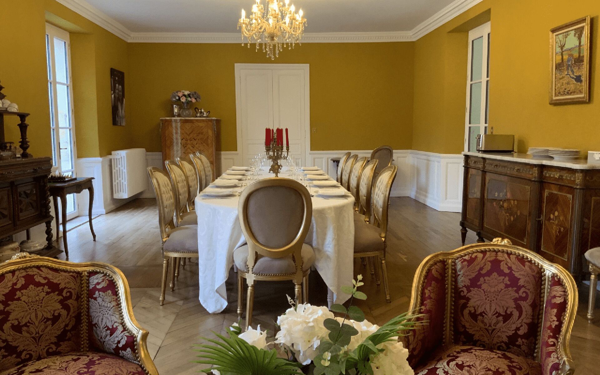 Une salle à manger élégante, avec une longue table dressée pour un repas, entourée de chaises richement décorées et surmontée d’un lustre.