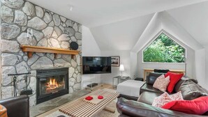 A cozy living room with a stone fireplace, a comfortable leather sofa, and a large window offering a view of greenery. - Golf course townhouse w/ free shuttle, parking (Whistler)