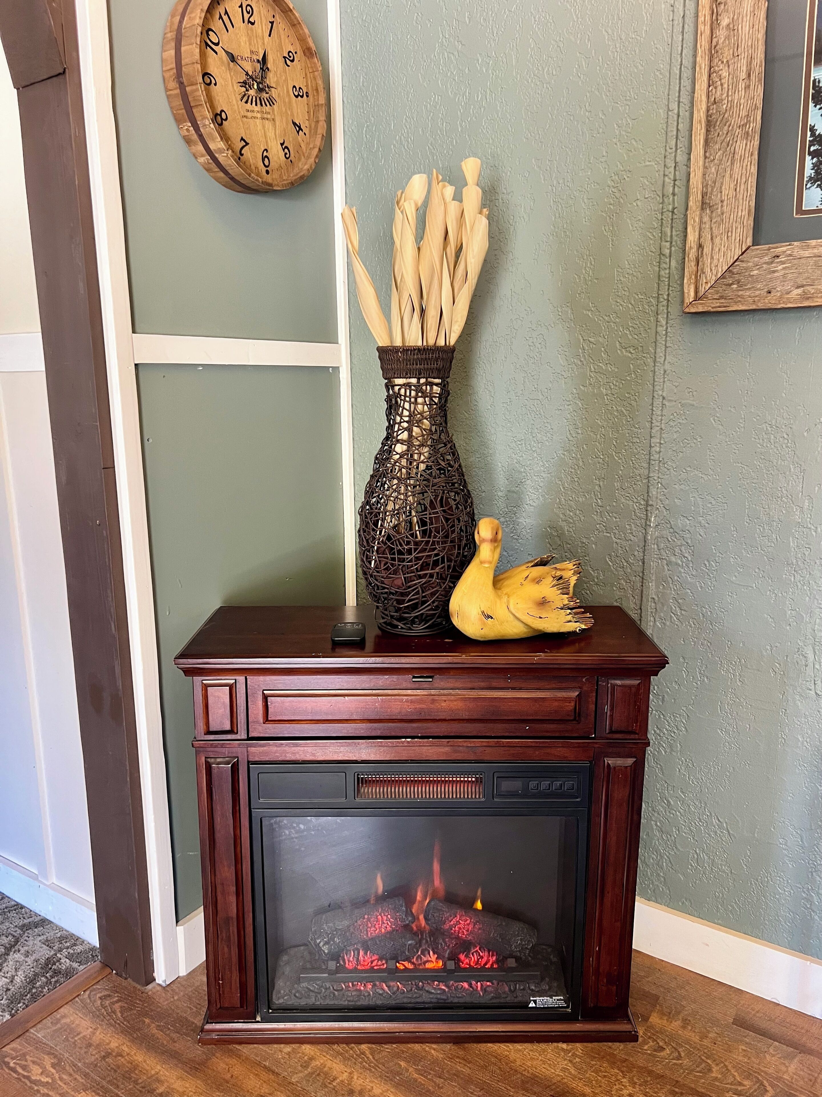 A fireplace with logs burning, a vase with dried plants, and a wall clock.