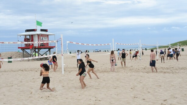Partido de voleibol de playa con jugadores y espectadores, una torre de salvavidas y una red.