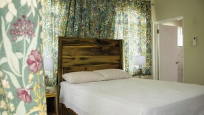 Un lit simple avec une tête de lit en bois, dans une pièce décorée de rideaux à motifs floraux et dont la porte se trouve sur le côté droit.