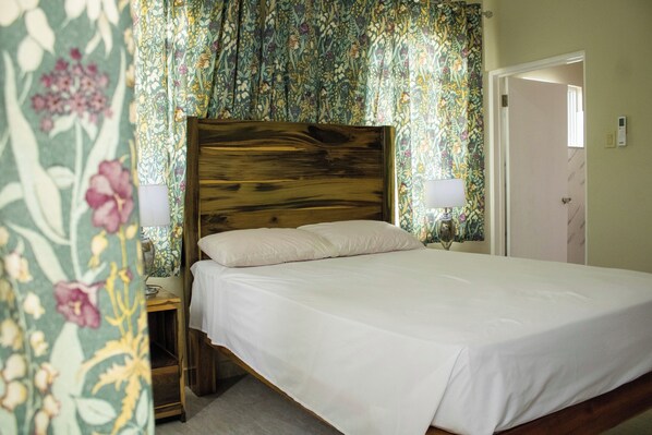 Un lit simple avec une tête de lit en bois, dans une pièce décorée de rideaux à motifs floraux et dont la porte se trouve sur le côté droit.