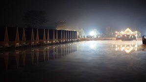 Vista nocturna de una propiedad frente al agua, con una fila de tiendas de campaña y un edificio cuyas luces se reflejan en el agua.