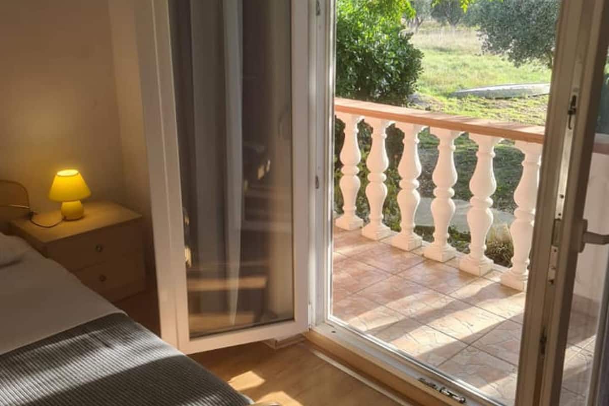 A bedroom with a bed, a bedside table with a lamp, and a sliding glass door leading to a balcony with a white balustrade.