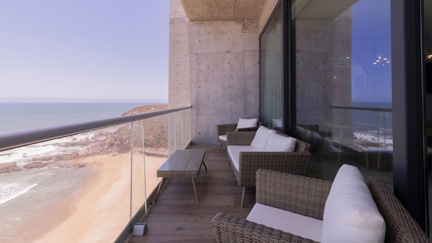 A balcony with wicker furniture and a glass railing overlooking a beach.