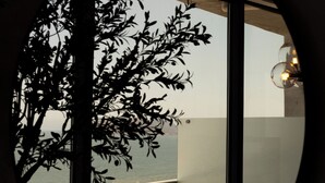 A view through a circular window overlooking a beach with waves, a wicker chair, and a lamp.