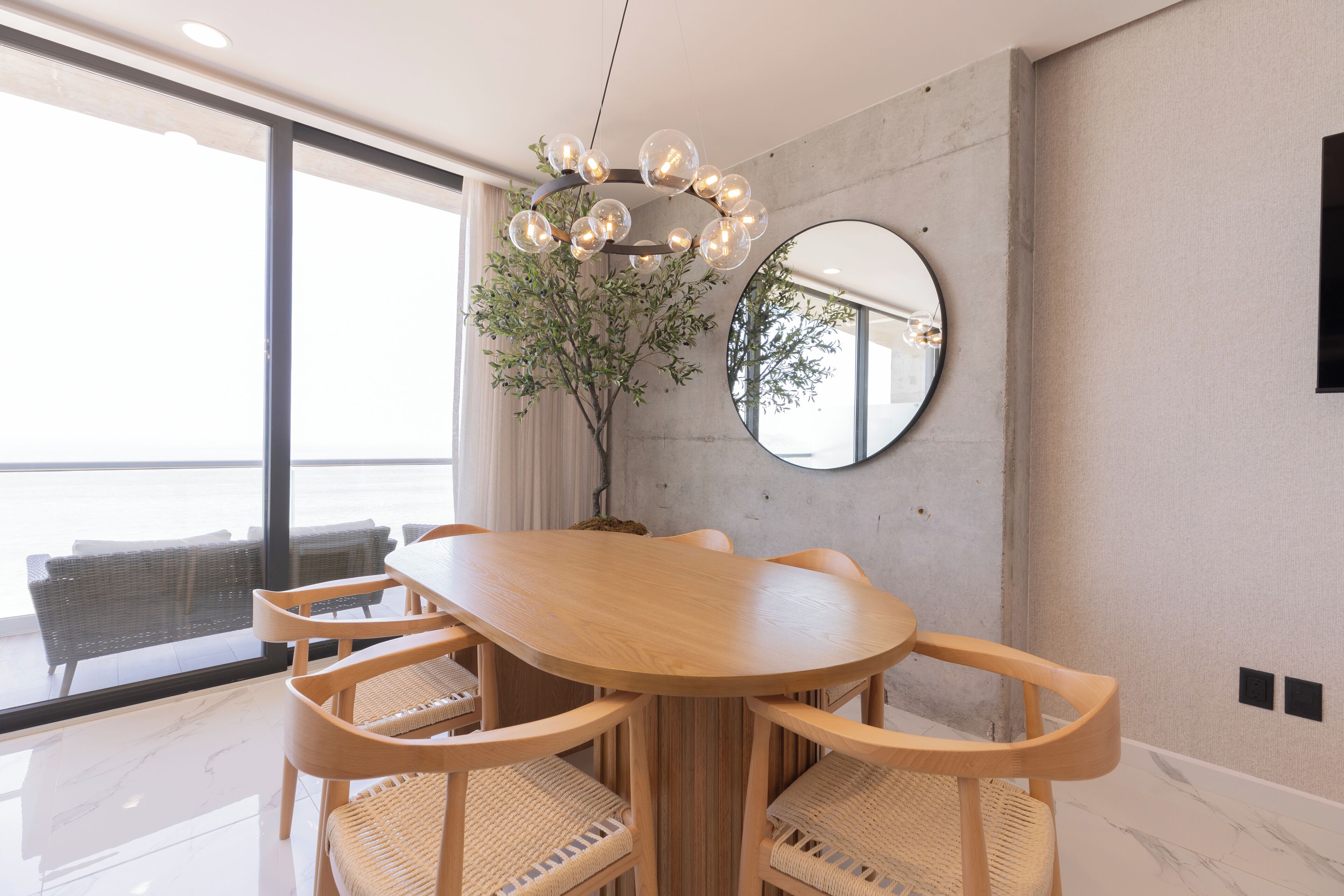 A modern dining area with a wooden table and chairs, a hanging light fixture, a potted plant, and a round mirror on the wall.