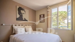 Une chambre à coucher avec un grand lit, une tête de lit dotée d’un luminaire, une œuvre d’art accrochée au mur, une citation inspirante et une fenêtre donnant sur des arbres.