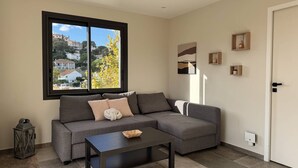 Un salon moderne avec un canapé d’angle, une table basse et une grande fenêtre donnant sur des immeubles et des arbres.