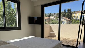 Une chambre à coucher avec un lit, un téléviseur fixé au mur et un balcon donnant sur le quartier.