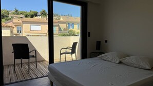 Une chambre à coucher comprenant un lit, deux chaises, une table de chevet et une grande porte-fenêtre coulissante donnant sur un balcon.
