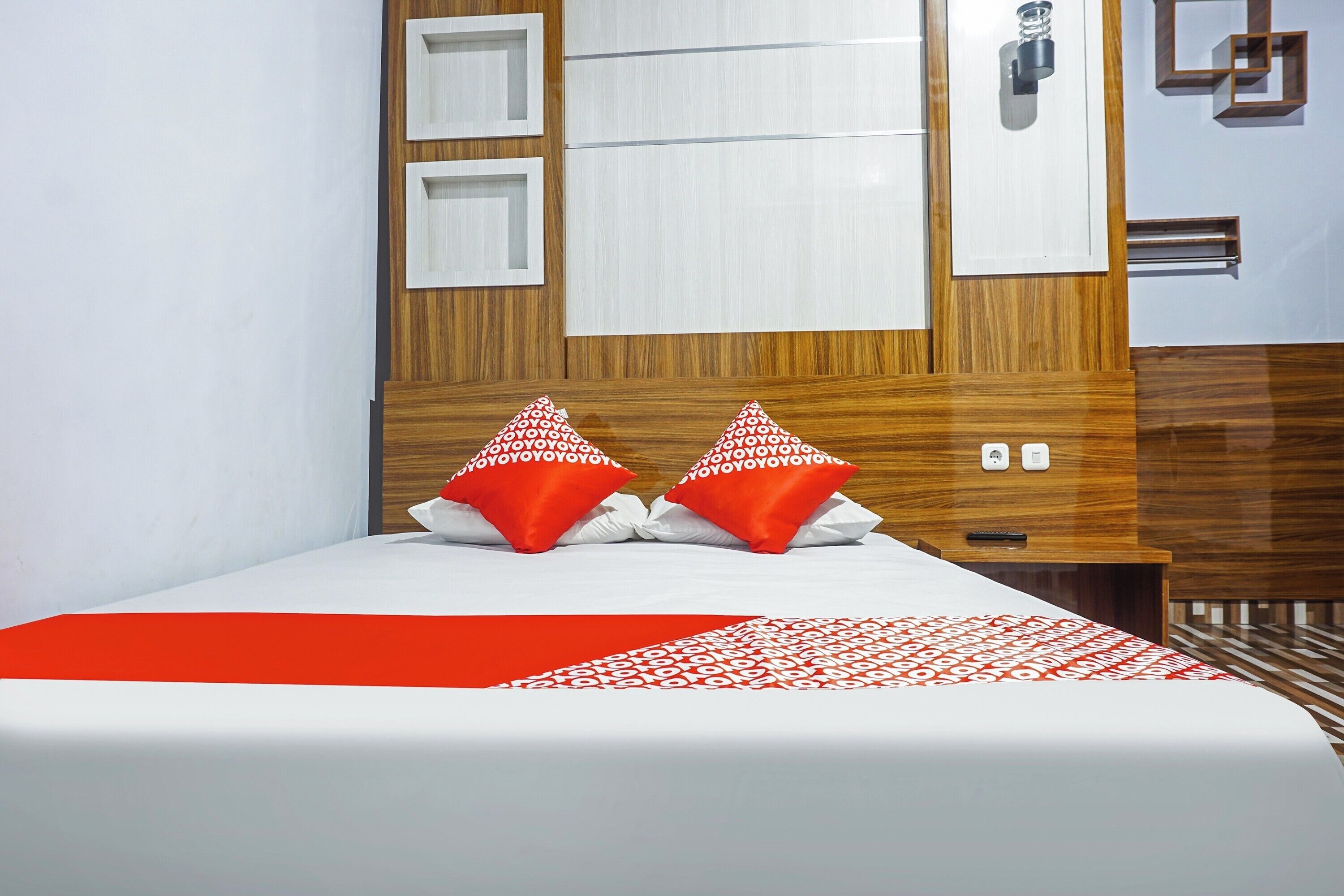 Una habitación de hotel con cabecera de madera, una cama con ropa blanca y roja, y una mesita de noche pequeña.