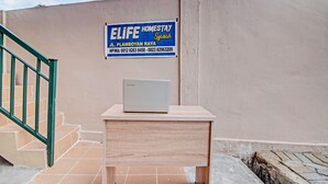 Ein Lenovo Laptop steht auf einem Holztisch vor einer beige farbenen Wand. Auf der Wand ist ein Schild zu sehen, das 'ELIFE HOMESTAY Syariah' bewirbt und Kontaktdaten enthält.