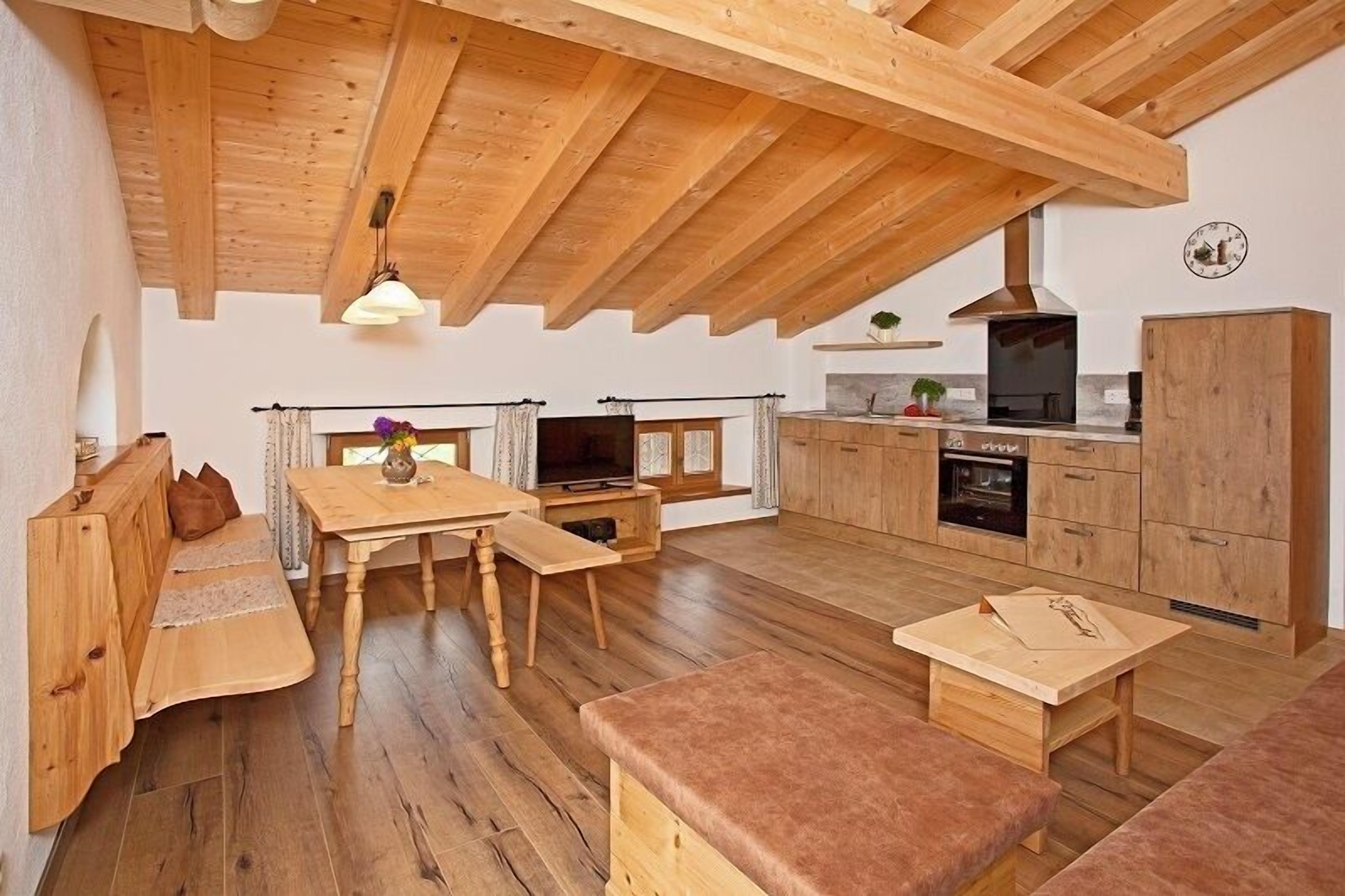 A cozy, open-plan living and dining area with wooden flooring and furniture, a built-in kitchen with modern appliances, and a sloped ceiling with exposed beams.