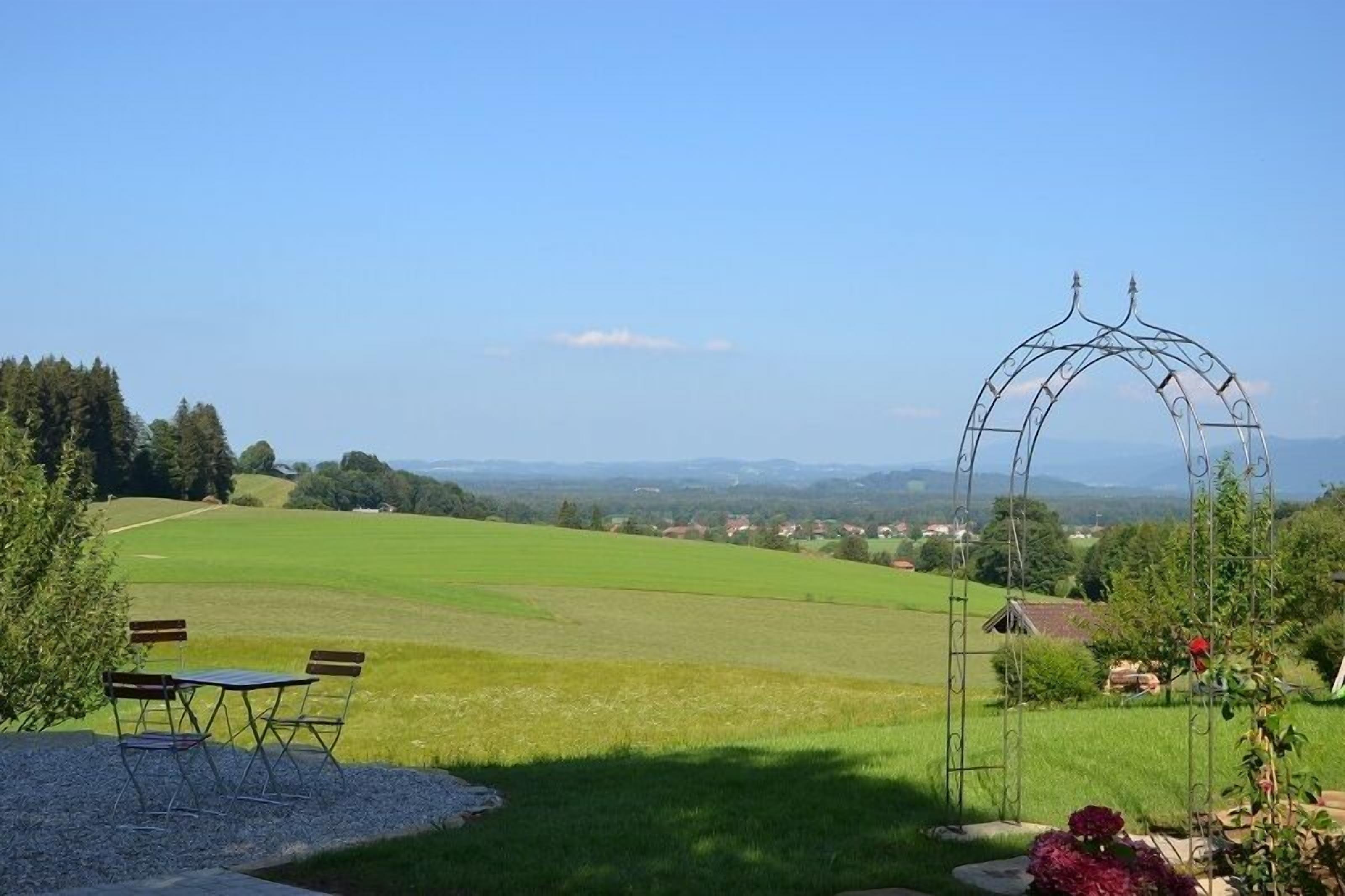 Un espace extérieur agréable, agrémenté d’une arche en métal, d’une table avec des chaises et offrant une vue sur un champ verdoyant et des maisons situées au loin.