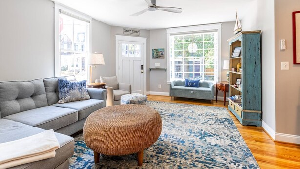 A living room with a grey sofa, a round ottoman, a blue patterned rug, a ceiling fan, and a bookshelf.