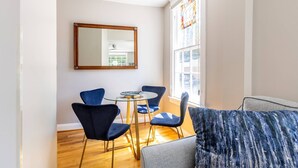 A modern living room with a glass table and blue chairs, a large window with a view, and a grey sofa.