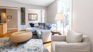 A modern living room with a sofa, ottoman, and lamp. There is a window providing natural light and a view of the outdoors.