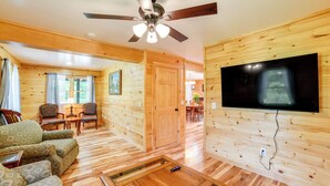 Interior acolhedor de uma cabana de madeira com piso de madeira, ventilador de teto, TV fixada na parede e um espaço de jantar com mesa e cadeiras.