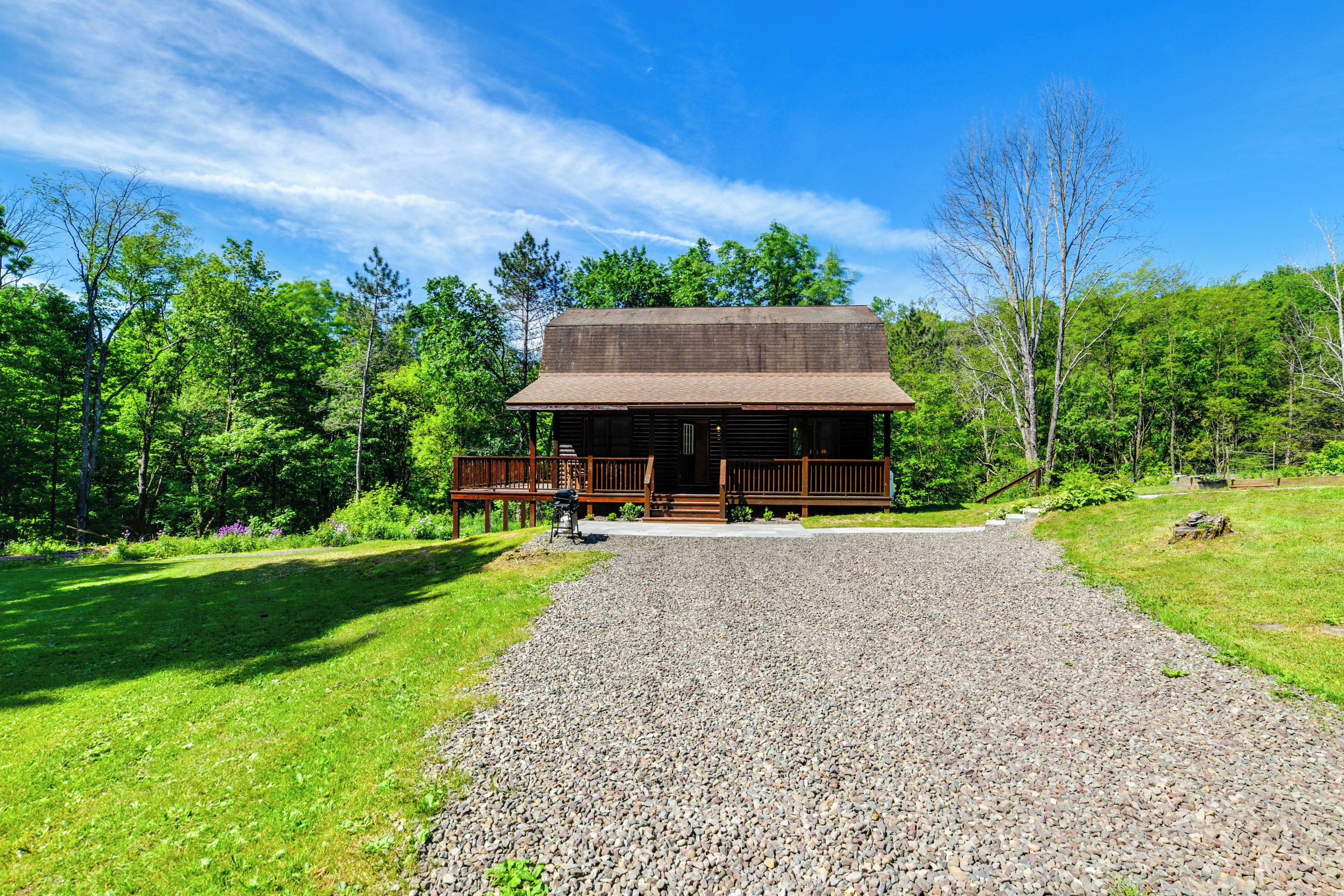 A rustic cabin with a gravel driveway leading to a wooden deck, surrounded by lush greenery.