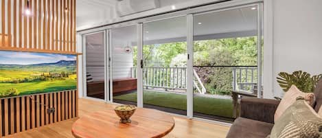 A modern living room with a wooden table, a sofa, and a large sliding glass door leading to a balcony with a view of greenery.