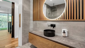 A modern bathroom with a wooden vanity, a round mirror, and a shower area visible through the glass door. - Bright on the River (Buffalo Apartment 1) (Bright)