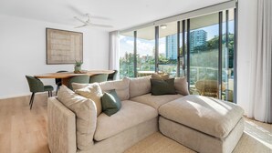 Un salon moderne avec un canapé d’angle, une table à manger avec des chaises et une grande porte-fenêtre coulissante donnant sur un balcon avec vue sur la ville.