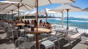 Un espace extérieur aménagé avec des tables et des chaises sous des parasols, offrant une vue sur une plage et l’océan.