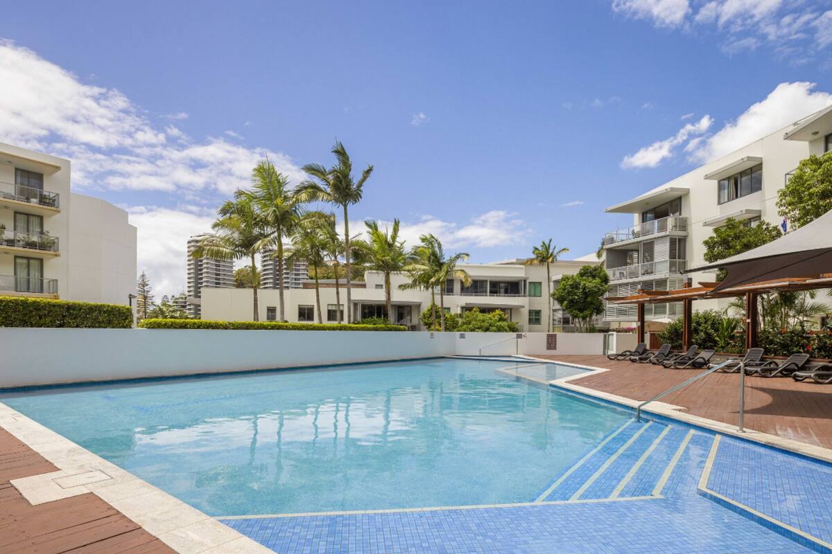 Una piscina con plataforma de baldosas, rodeada de edificios modernos con balcones y palmeras.