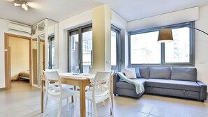 A modern living room with a sofa, a dining table with chairs, and large windows. - GuestHost - Sebeto Apartment x3 with Parking & Netfilx (Napoli)