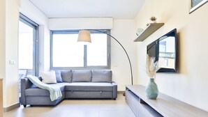 A modern living room with a grey sectional sofa, a floor lamp, a flat-screen TV mounted on the wall, and a small plant on a wooden console table. - GuestHost - Sebeto Apartment x3 with Parking & Netfilx (Napoli)