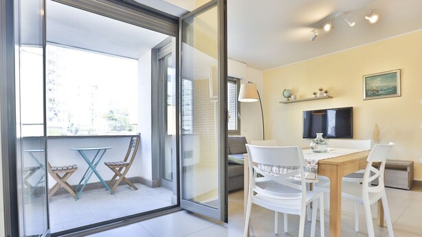 A modern living room with a glass door leading to a balcony, a dining area with white chairs, a TV on the wall, and a small shelf with decorative items. - GuestHost - Sebeto Apartment x3 with Parking & Netfilx (Napoli)