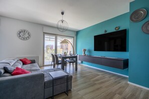 A modern living room with a grey sofa, red cushions, a dining area with chairs, a TV mounted on a teal wall, and a large window.