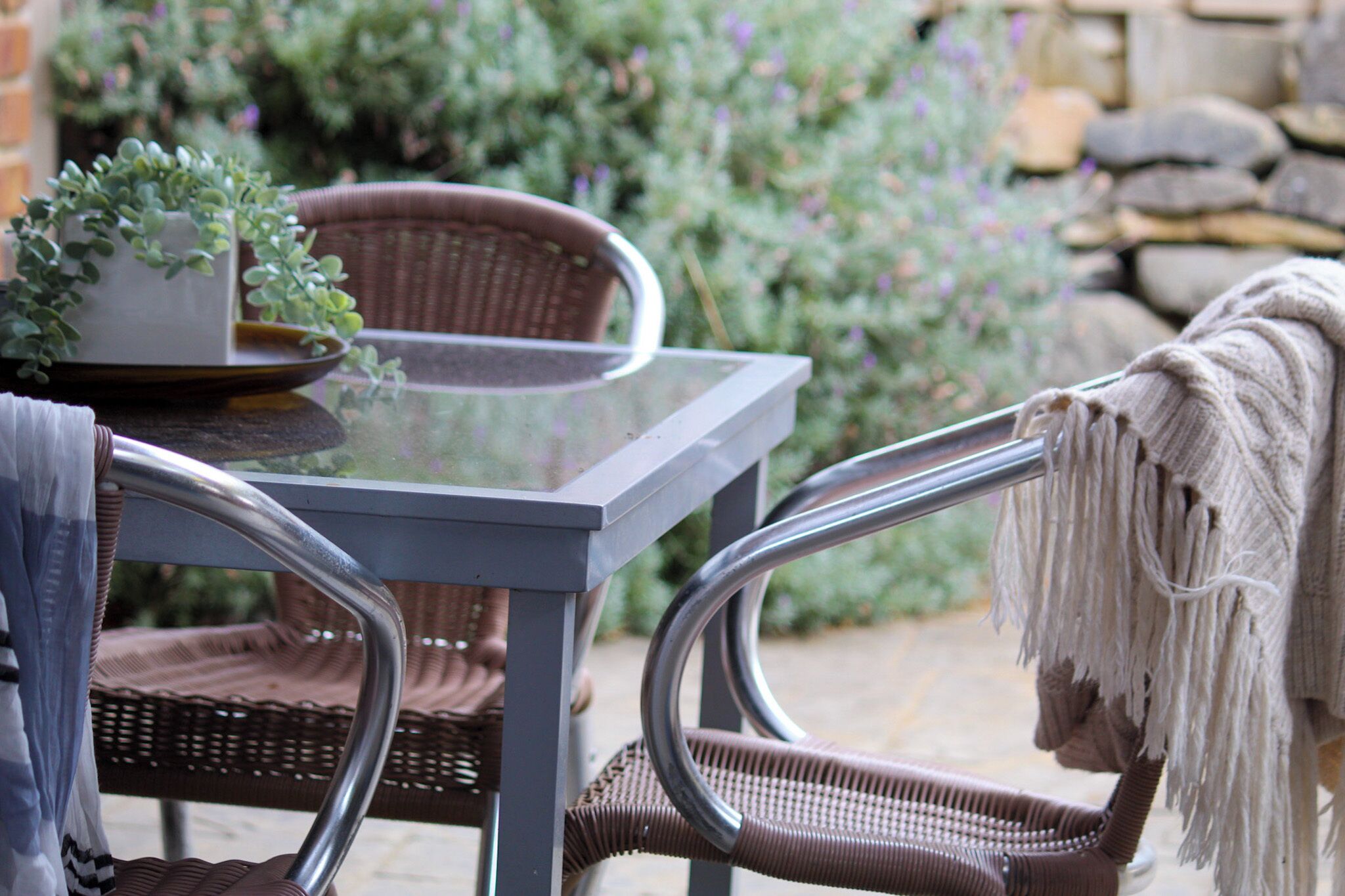 A glass-top table with wicker chairs, a potted plant, and a stone wall in the background.