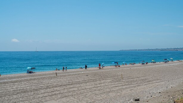Una spiaggia con persone, ombrelloni e una barca a vela all'orizzonte.