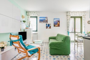 Quarto com sofá verde, cadeira de madeira, mesa pequena, televisão e radiador.